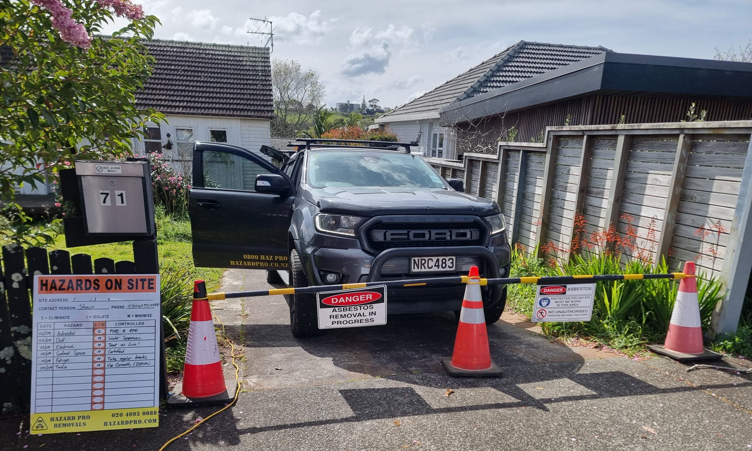 Site photo with hazard boards identifying asbestos removal site boundary. Hazard Pro service vehicles covering Auckland, Hamilton, Tauranga, Matamata, Gisborne, Taupo and every town in between.