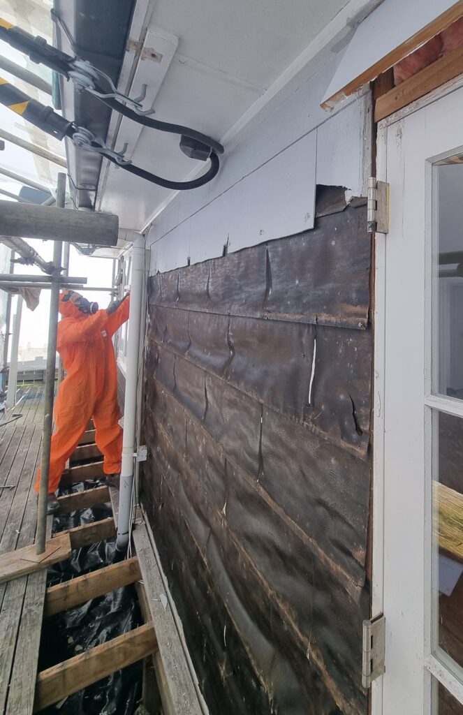 Certified asbestos removal technician dismantling exterior cladding in protective gear.