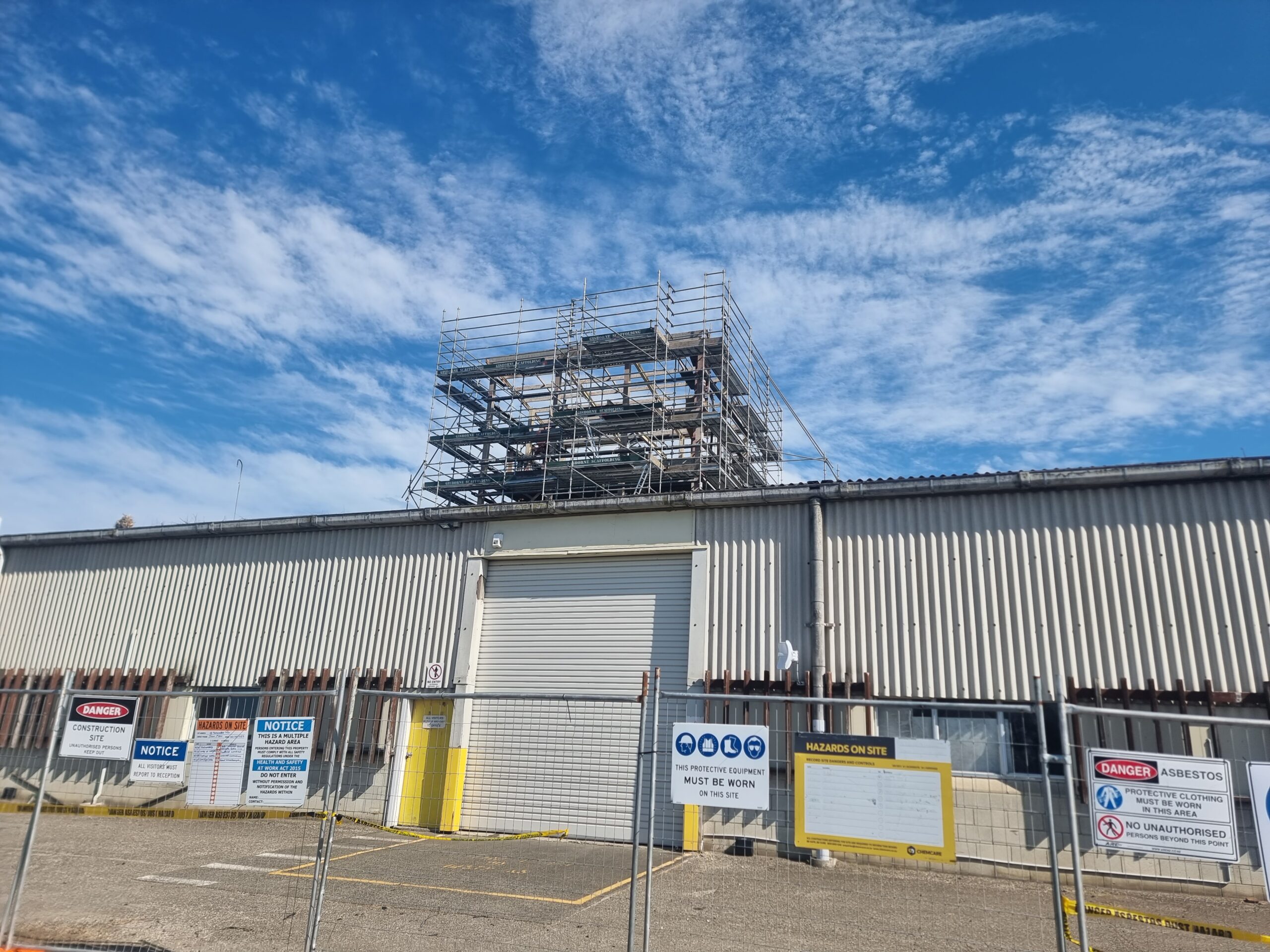 Commercial warehouse with Super6 asbestos cladding, asbestos roof sheets, and fibre cement gutters prior to removal, with tower cladding and roofing already dismantled in the background. Hazard Pro signage and barriers indicate a safe asbestos removal site.