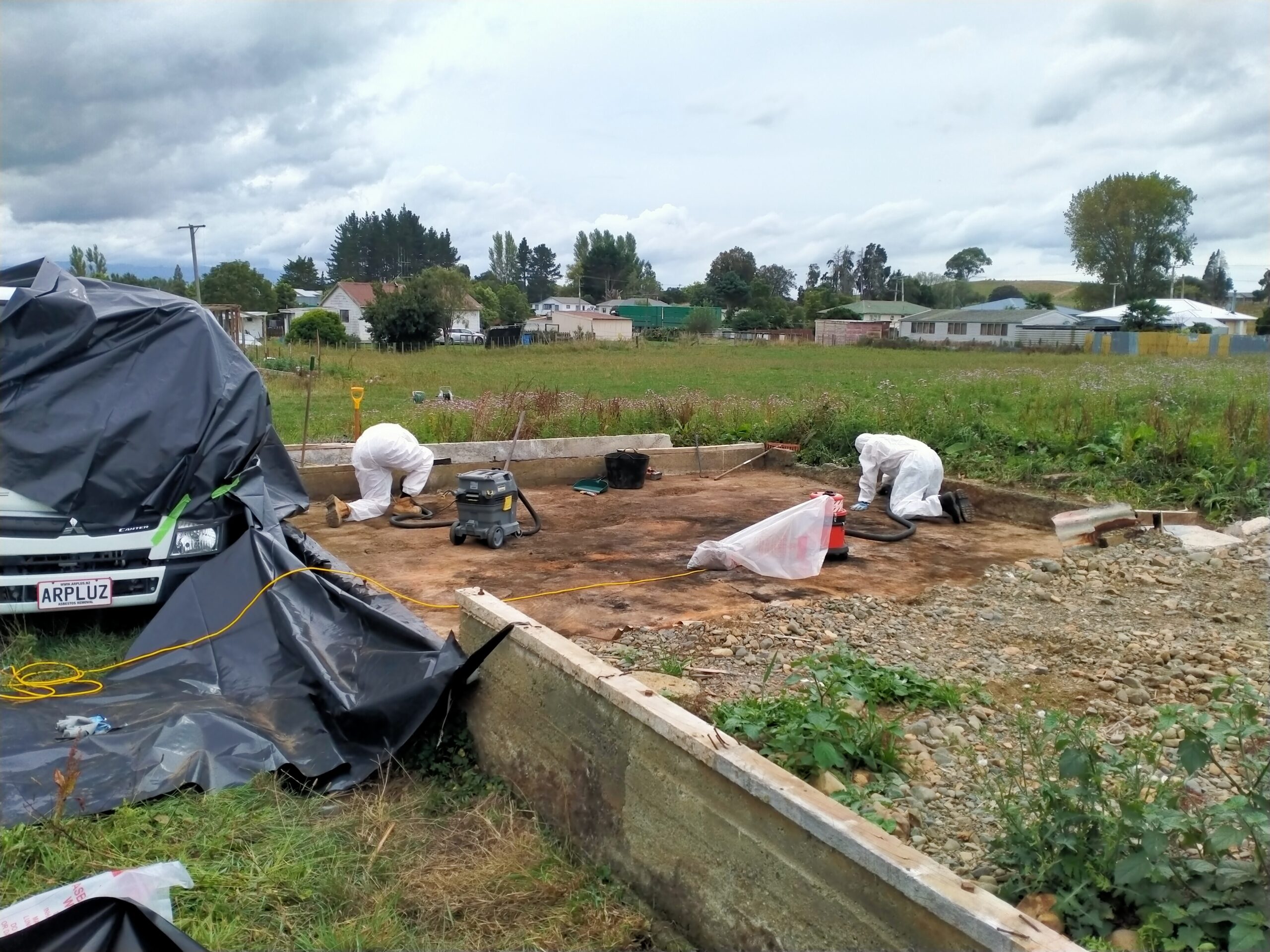 Two Hazard Pro asbestos removal professionals in full protective gear using HEPA vacuum equipment beside a vehicle wrapped in protective poly sheeting during soil remediation at a contaminated site.